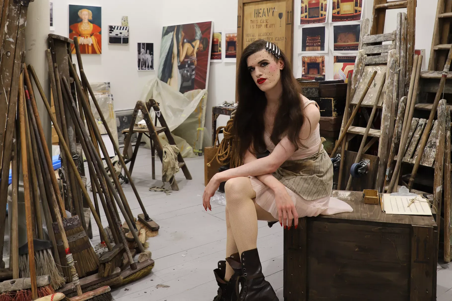 Very butch appearing woman sitting with arms crossed on knees. Long brown hair, heavy makeup, big black boots. In an art studio.