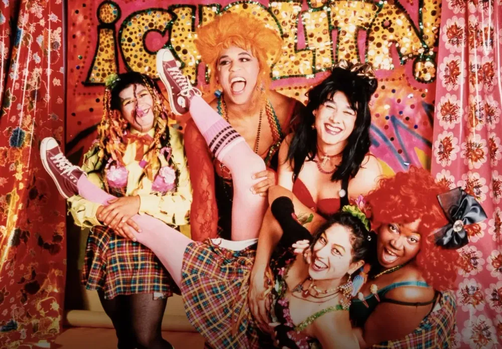 Colorful photo of several people with very happy expressions who are dressed in elaborate costumes and wigs. The mood is lively and energetic.
