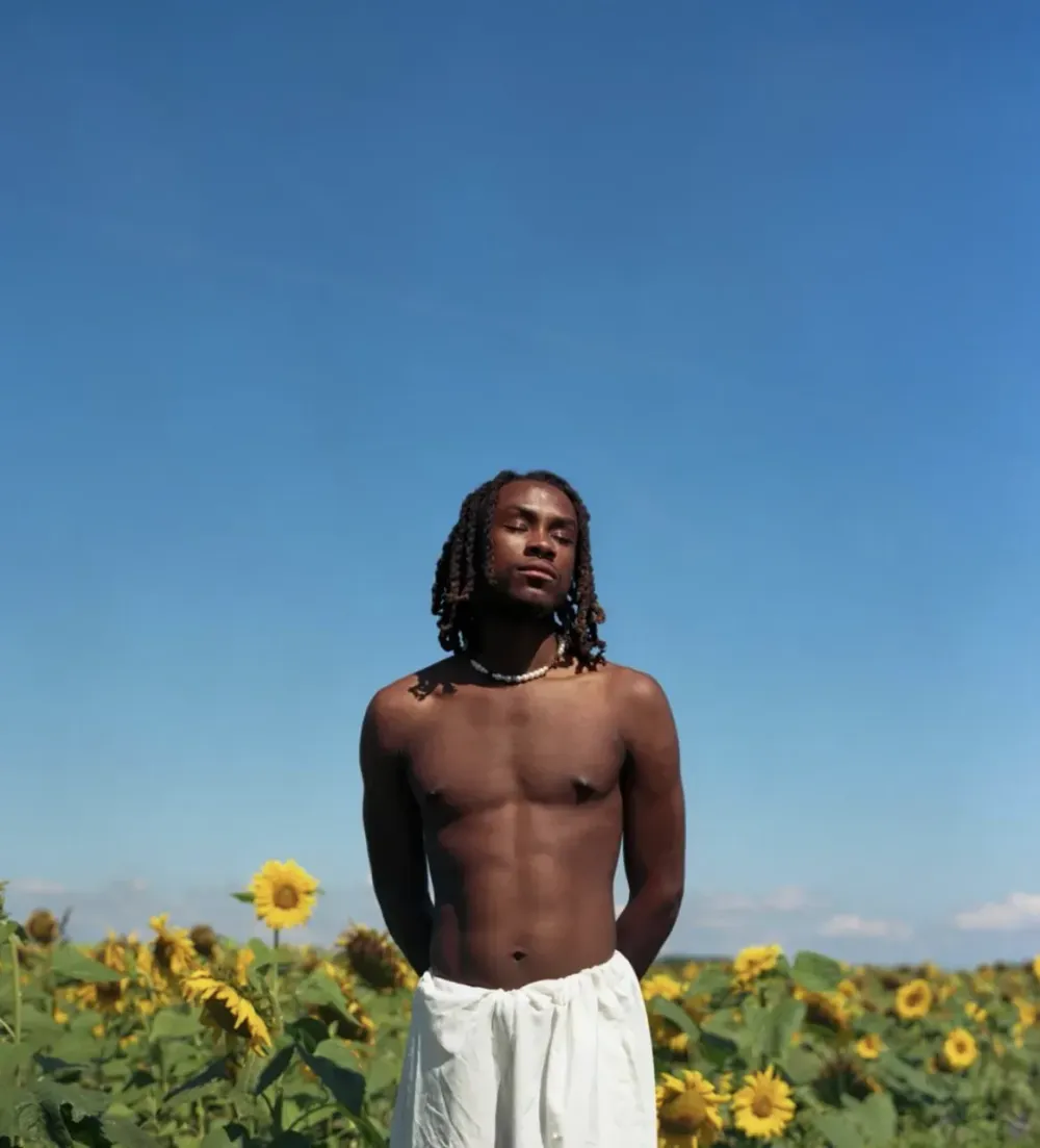 Colorful photograph of a young black man with shoulder-length braided hair. He is shirtless, with closed eyes, standing in front of a field of sunflowers.