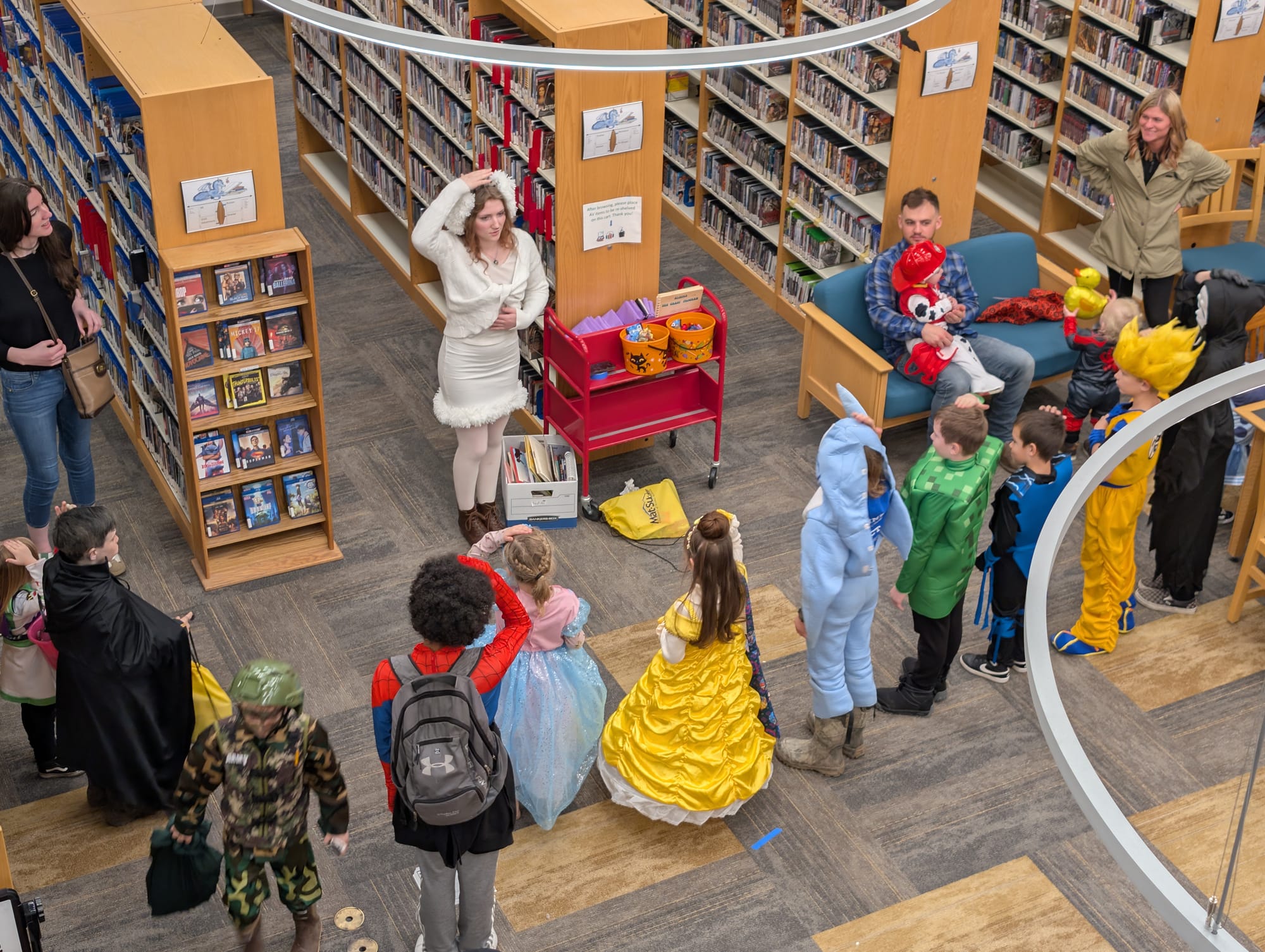 A woman in a white dress leads a group of six or seven kids in Simon Says in a library.