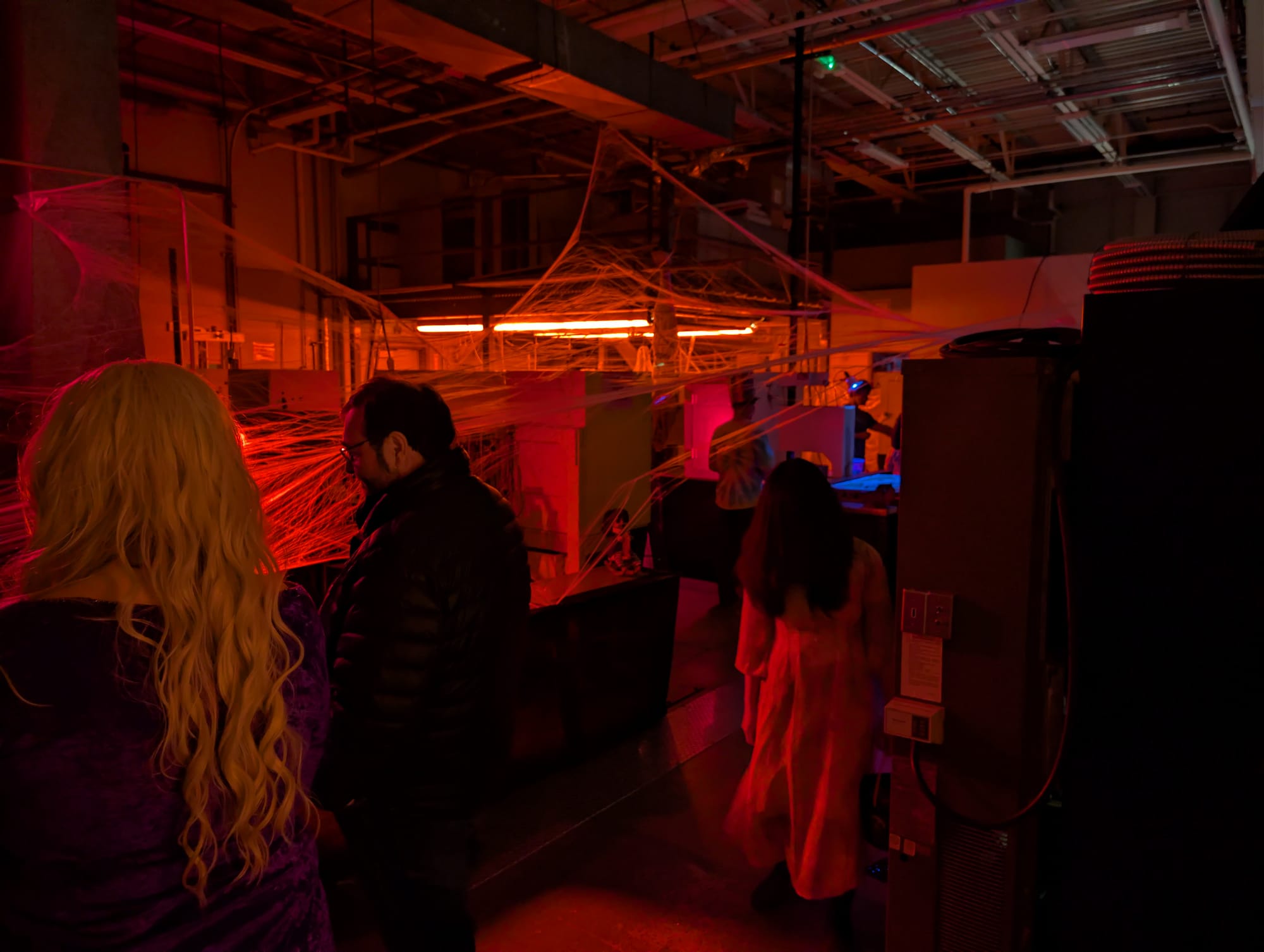 Four people look at animatronics inside a room lit with red light and decorated with cobwebs.