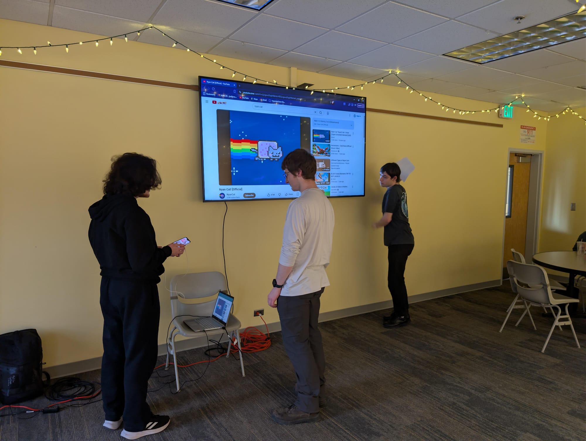 Three students standing in front of a laptop attached to a TV. There are cords visible running between TV and laptop.