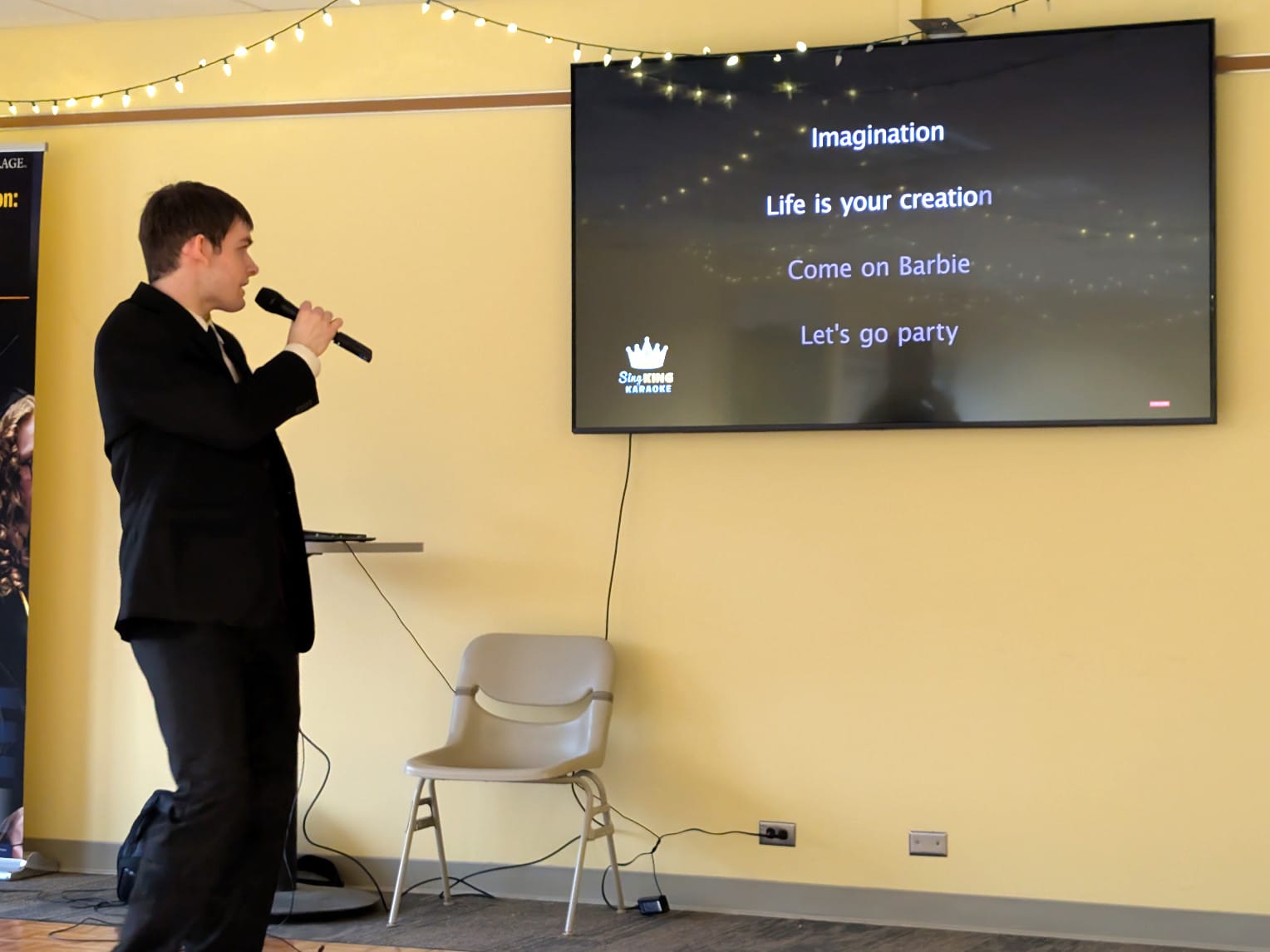A man in a dress suit holds a microphone and look sat a monitor that displays the lyrics to Aqua's Barbie Girl.