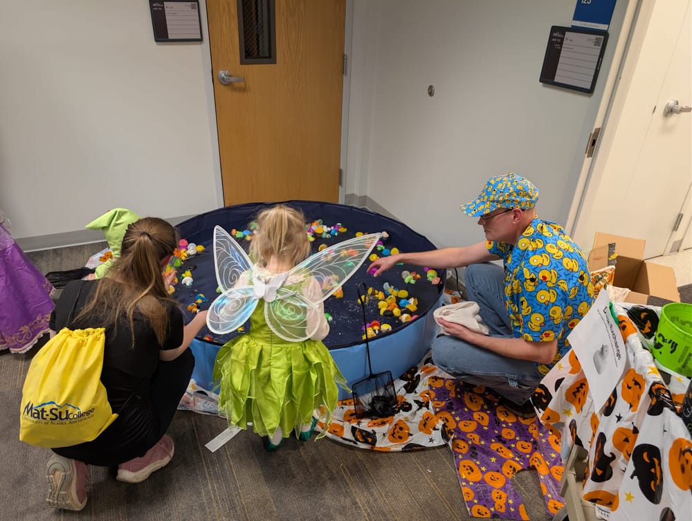 A man in a shirt with many ducks printed on it squats next to a small pool. A child is scooping out ducks with a net.