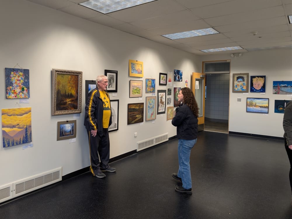 A woman speaks to a man in the middle of an art gallery. They are flank by art on every wall.