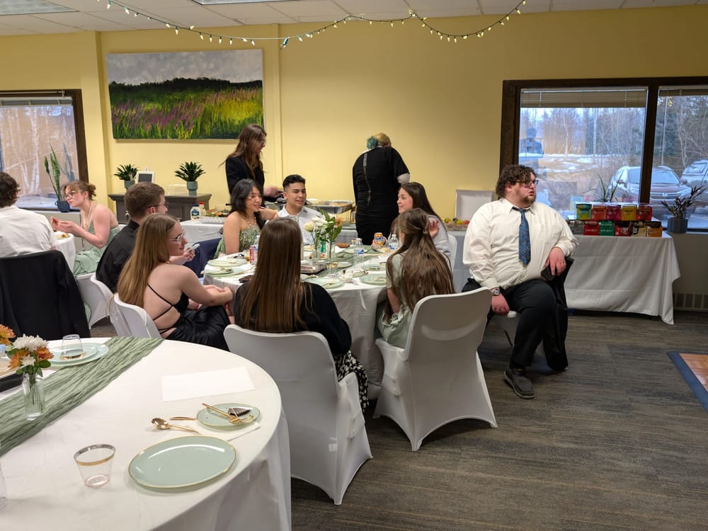 Students in formal attire sitting around a table. Students coverse or look at the dance floor.