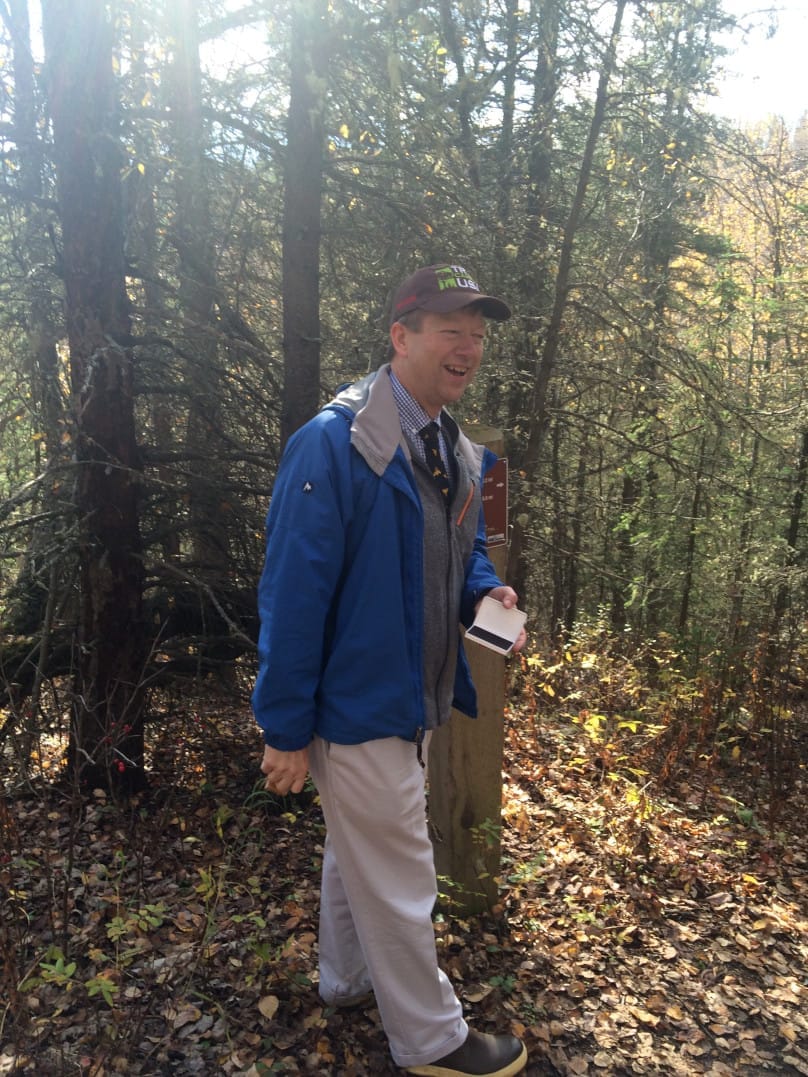 A smiling man stands with a stamp in a wooded scene. Autumnal leaves litter the ground.