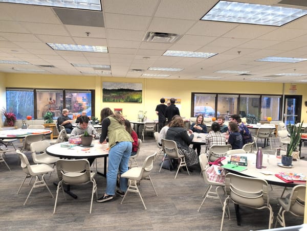 A photo of the campus cafeteria. Students and employees sit at three tables, happily playing games or eating. Two students stand at a table of food.