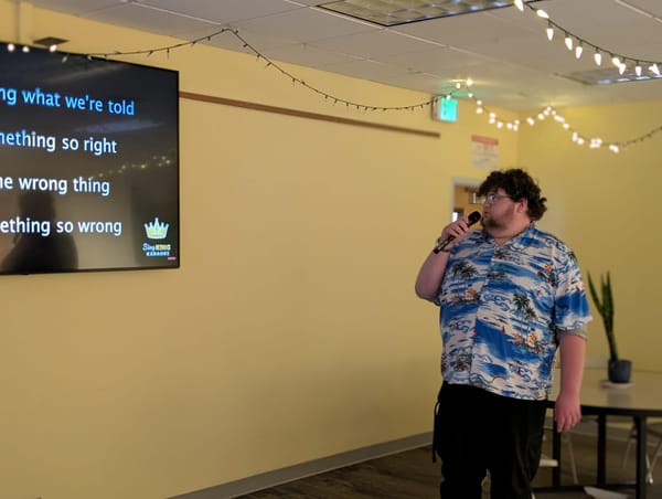 A student in a Hawaiian shirt looks at a TV displaying karaoke prompts. He has a microphone in hand.