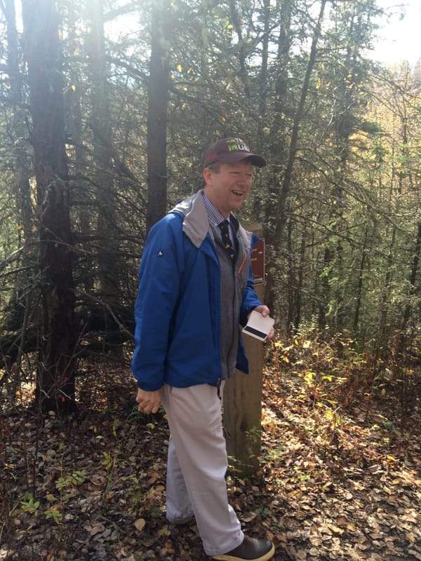 A smiling man stands with a stamp in a wooded scene. Autumnal leaves litter the ground.