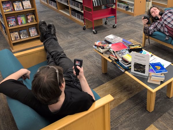 Two students, reclining on couches, watch an Angela Lansbury fitness program on small TVs.