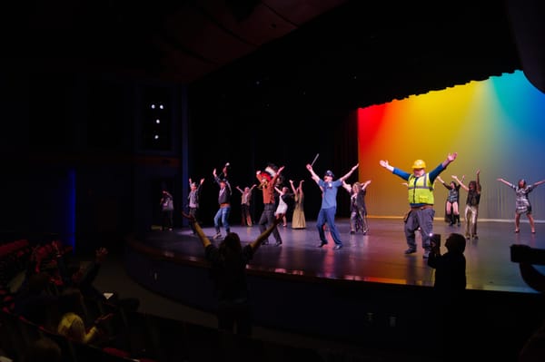 A theater stage with several people dressed in costumes dancing in the foreground. Behind them are other dancers. The first couple of rows of the audience can be seen.