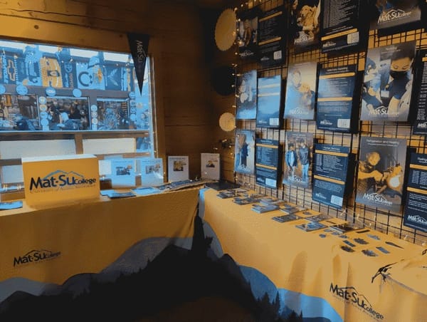 Interior of the Mat-Su College cabin at the Alaska State Fair. Tables are lined with gold and blue tablecloths, on which sit free handouts and brochures.