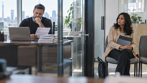 A hiring manager reviews applications while an experienced job seeker waits on the opposite side of a glass office partition.