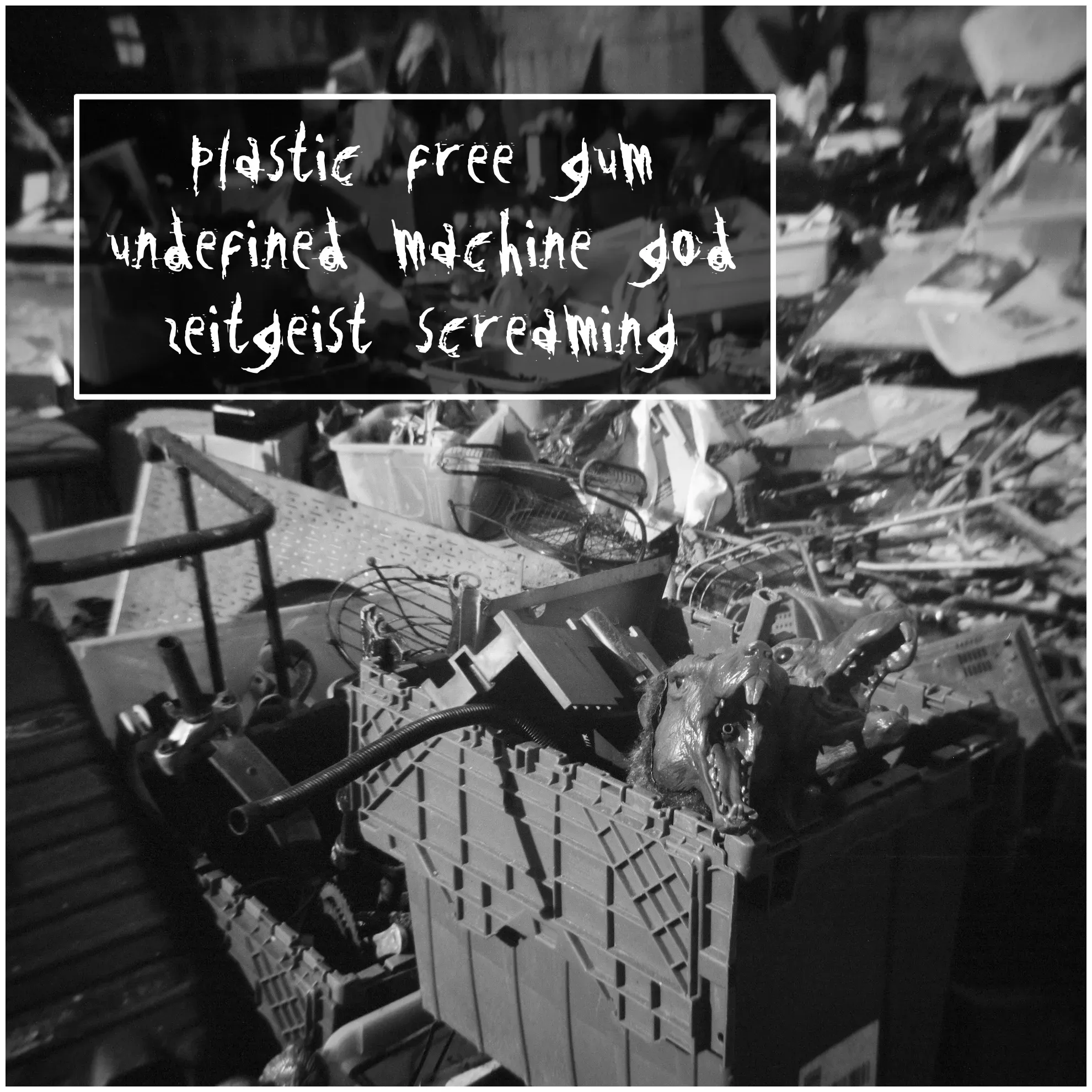 Square black and white photo of a huge pile of random junk on a sidewalk. In the foreground are two plastic dog heads with wide open jaws and jagged teeth. The words, “plastic free gum, undefined machine god, zeitgeist screaming,” are superimposed.