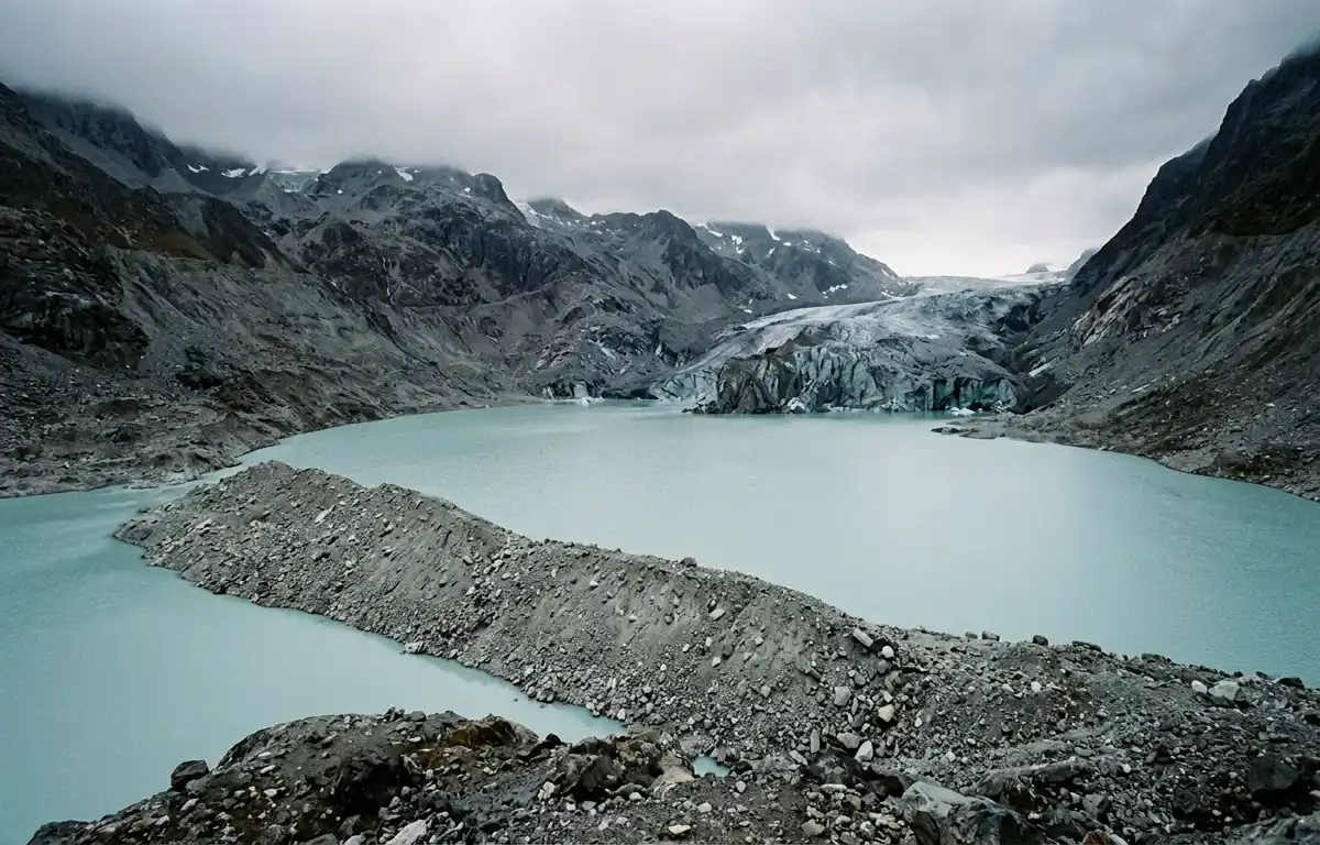 A high-altitude glacial lake of pale turquoise meltwater in flat overcast light, with a retreating glacier at the far end and a long ridge of loose rock and gravel debris in the foreground. 