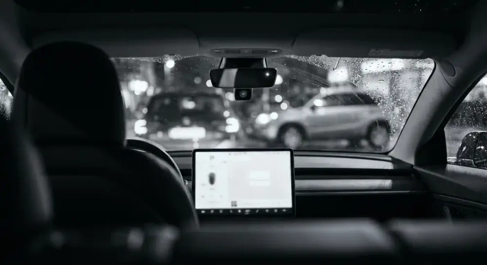 The dark interior of an empty car at night, its dashboard screen glowing and a small camera lens visible near the rear-view mirror.