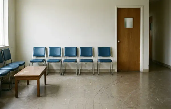 An empty medical waiting room in flat natural daylight, with a row of chairs against a pale wall and a closed door. No people, signage or text are visible.