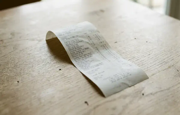 A close-up of a printed paper receipt on a pale wooden table in flat natural light, showing columns of small unreadable figures. No text or branding is legible.