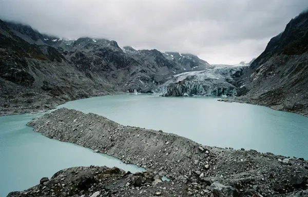 A high-altitude glacial lake of pale turquoise meltwater in flat overcast light, with a retreating glacier at the far end and a long ridge of loose rock and gravel debris in the foreground. 