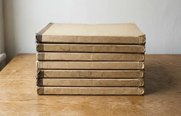 A stack of closed buff-coloured case folders on a plain wooden desk in flat daylight, with slightly worn covers and nothing else on the desk. No people, text or labels are visible.
