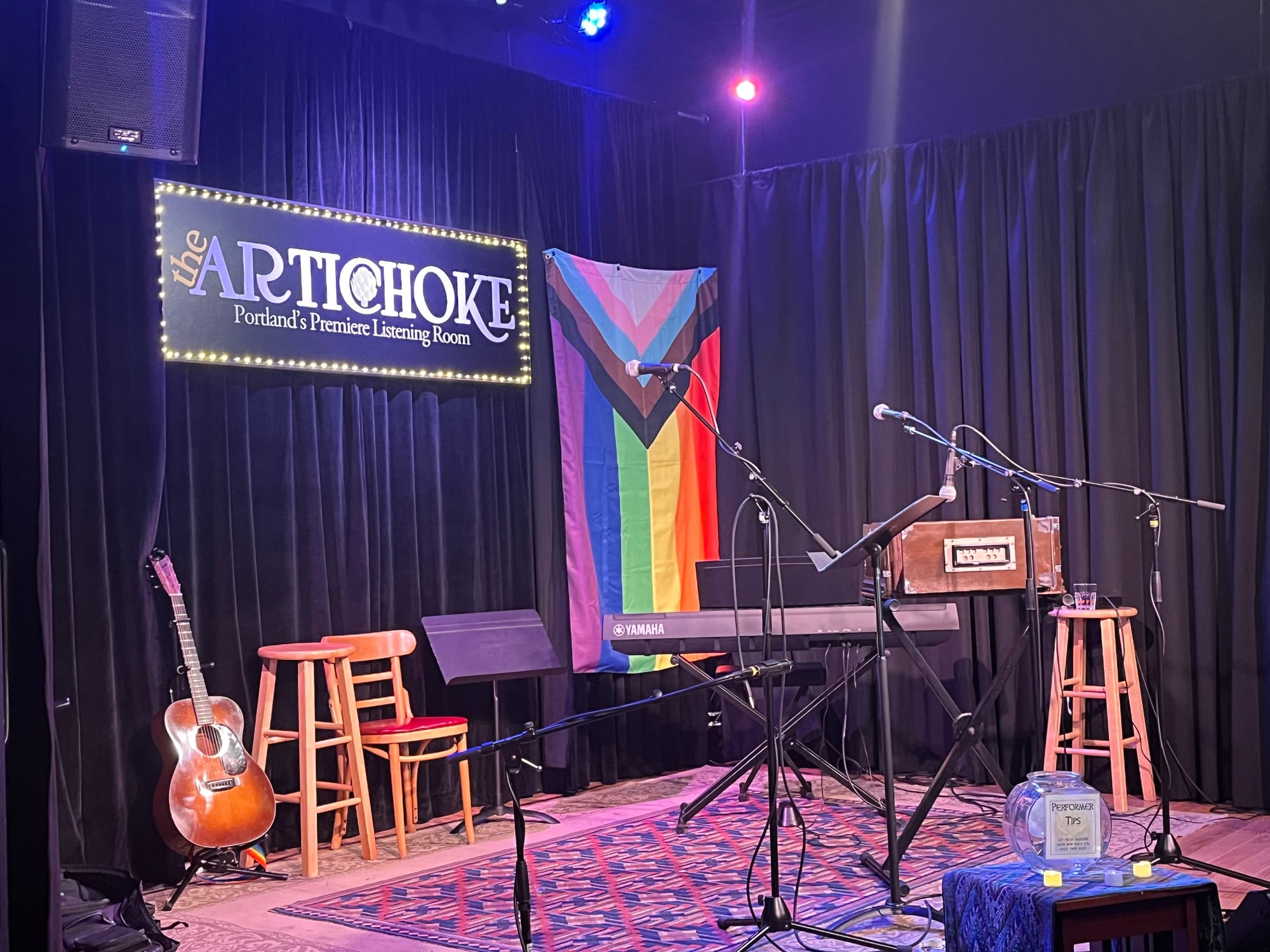 A small stage with a guitar and keyboard and a pride flag in the background. A sign says “the Artichoke: Portland’s Premiere Listening Room”