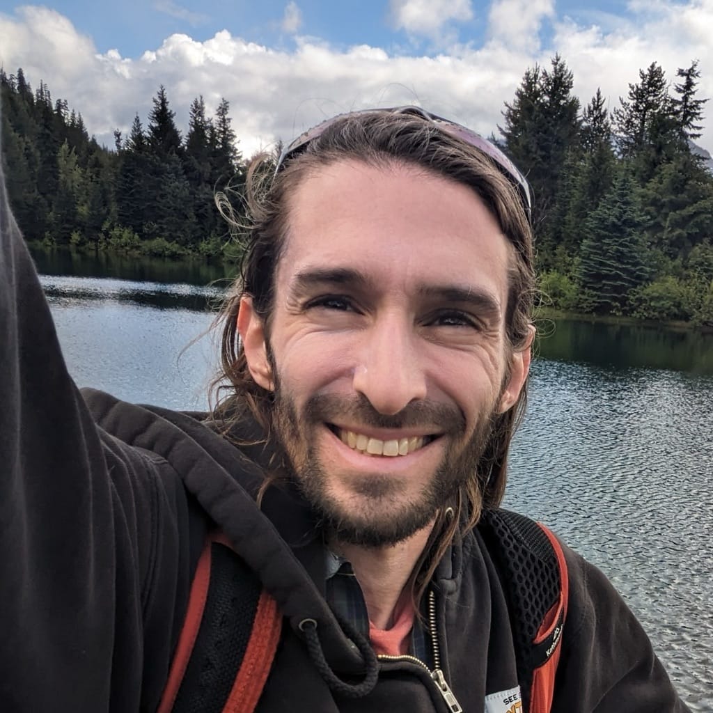 A bearded and smiling white man with long brown hair with a lake, trees, and a cloudy sky in the background