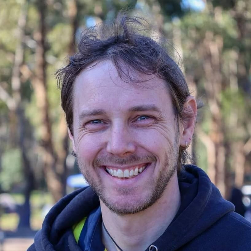A masc-presenting bearded white person with windswept hair smiling broadly at the camera, and out-of-focus trees in the background