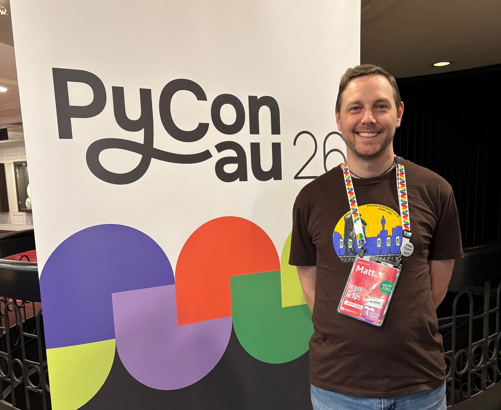 A smiling person with a brown shirt and conference lanyard stands next to a sign or PyConAU 2026.