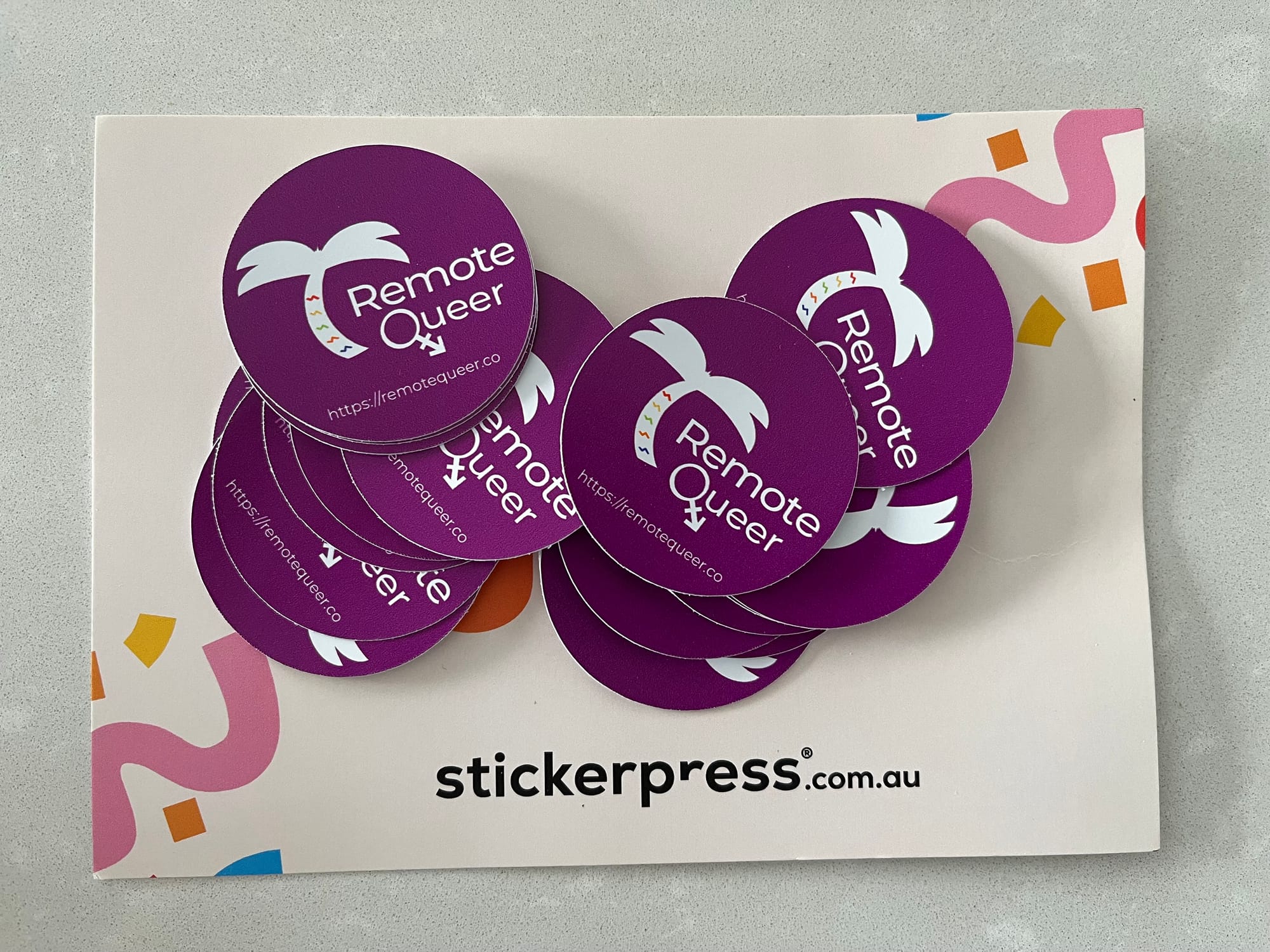 Around a dozen round, purple stickers with the Remote Queer logo, atop a paper that says stickerpress.com.au