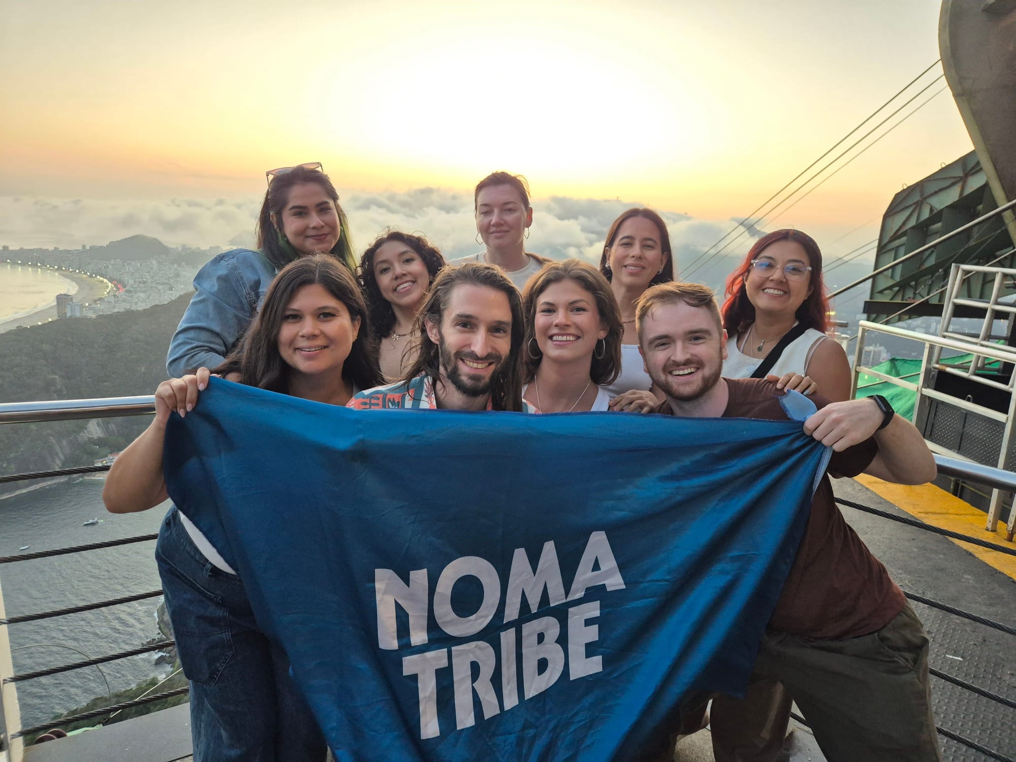 Ten smiling people on top of a viewpoint overlooking a city, holding a blue flag that reads “NOMATRIBE”