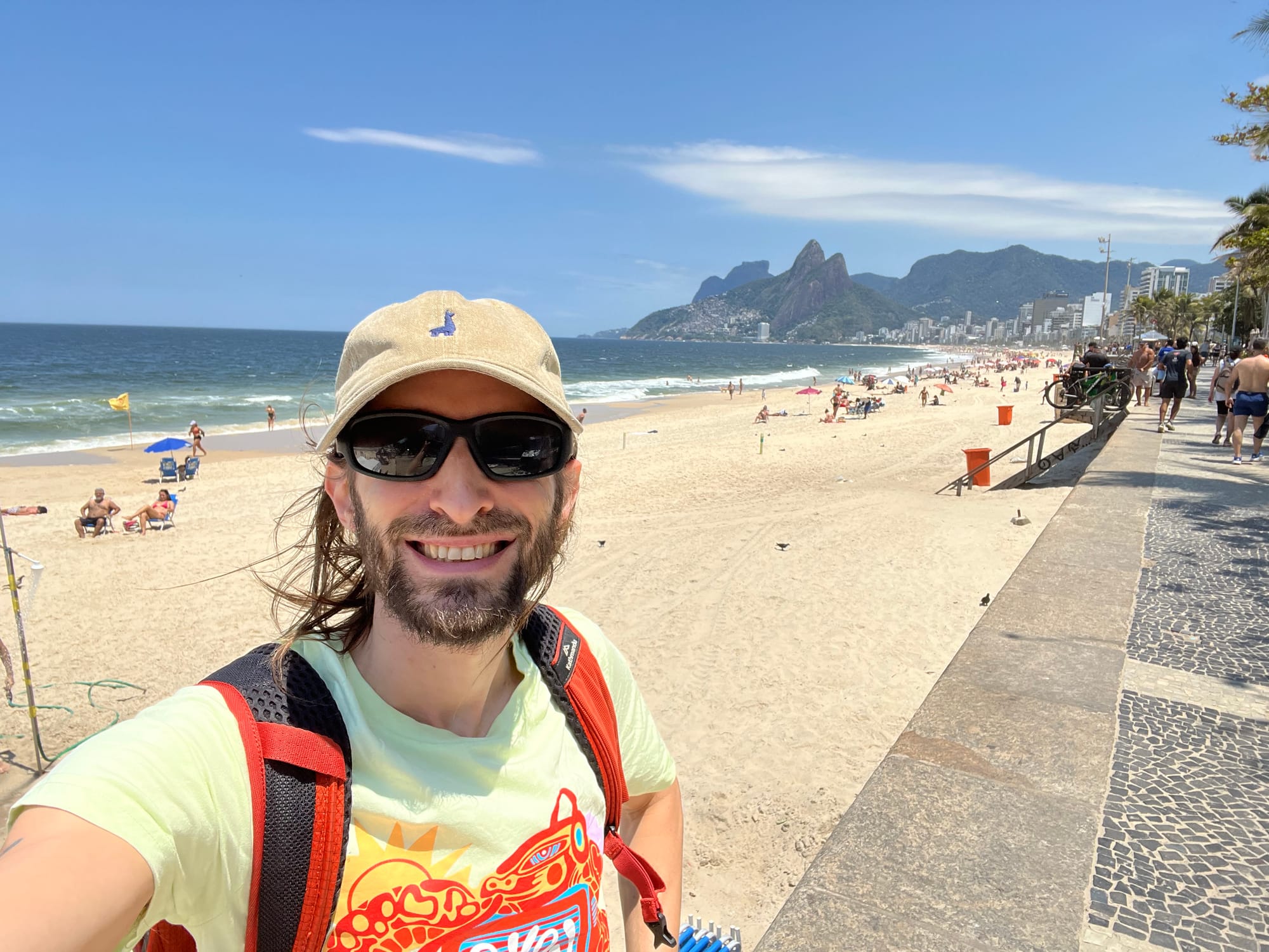 A guy with a hat, sunglasses, and backpack smiling in front of a sandy beach with sharp hills in the distance.