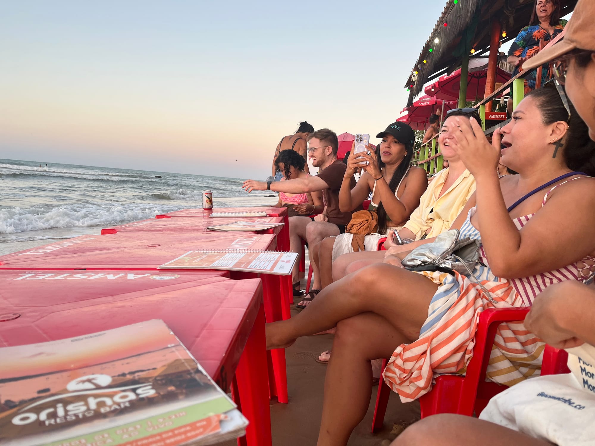 A group of people sitting at red tables right next to the ocean as if the tide is going to sweep them away.