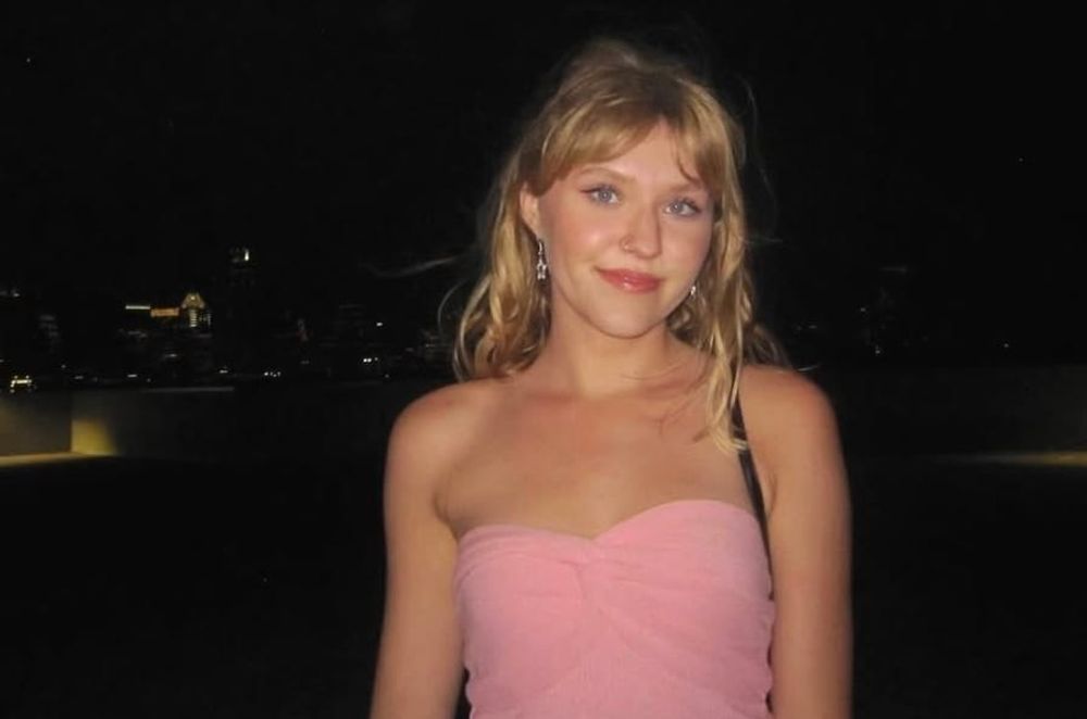 Blonde woman in pink sleeveless shirt stands in a dark parking lot at night. She is illuminated by flash.