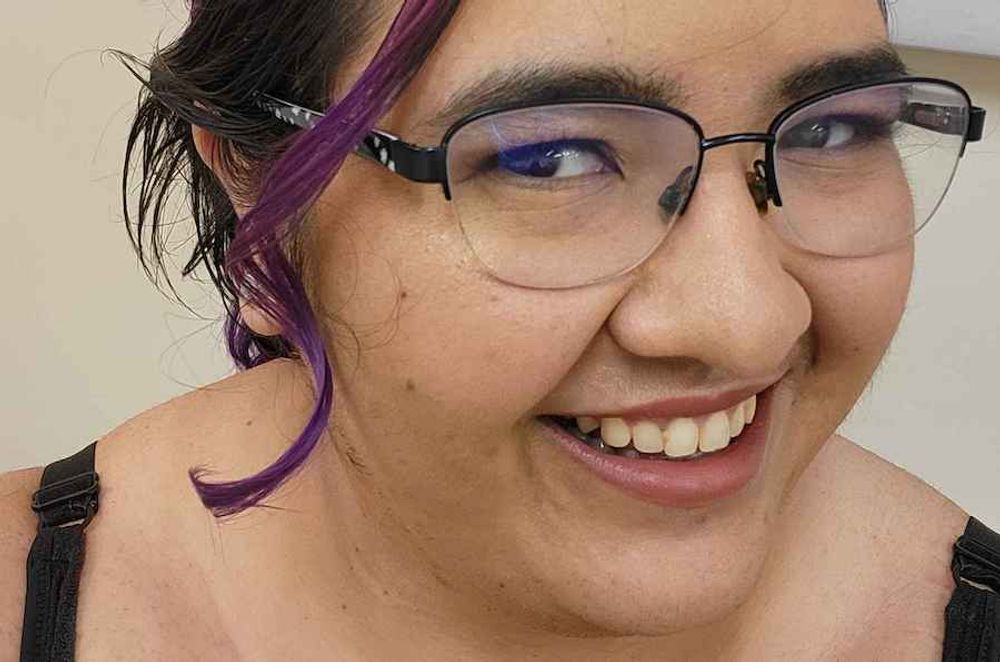 A young woman wearing glasses,  with purple hair tied up in a bun. She is smiling and her head is at a 3/4 angle.