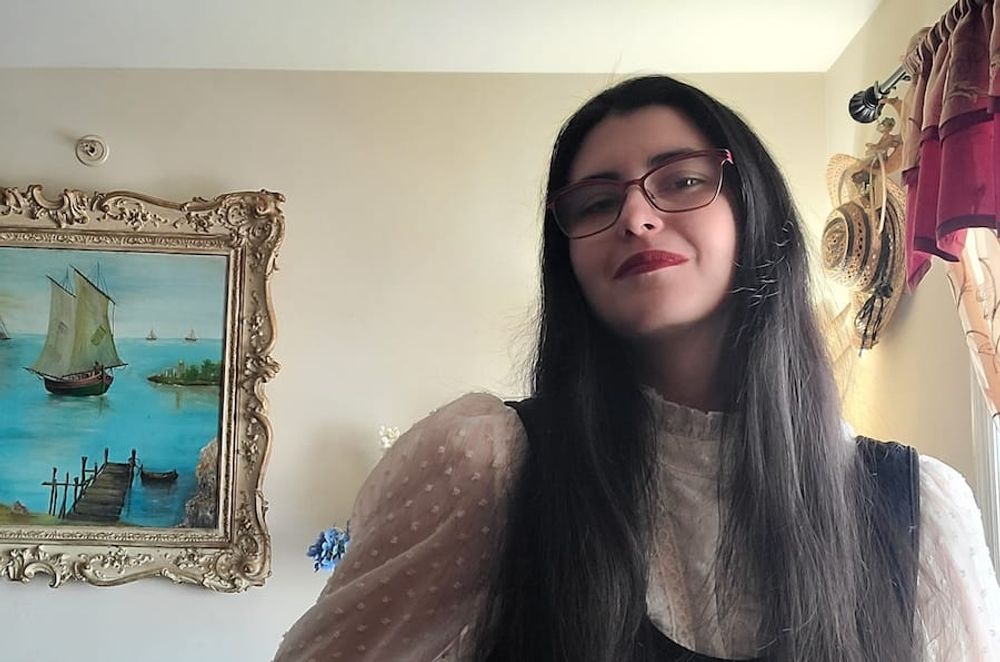 The poet stands in a black dress with a white blouse in front of a painting of a port in Italy, by her great-great aunt, Gigetta. Her hair is long and she is wearing red glasses and lipstick.