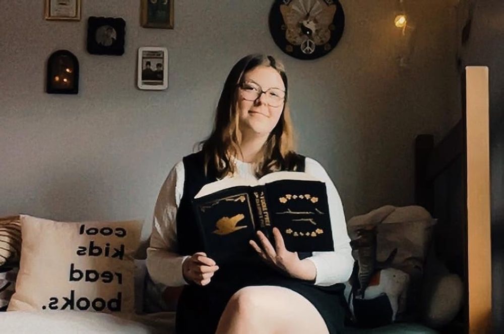 Courtney sits on a bed with a book open in hand, looking and smiling at the camera.
