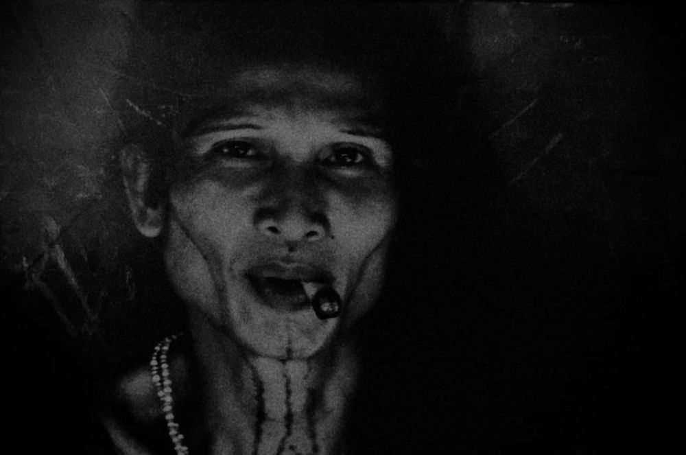 Grainy and dark black and white photo of an androgynous person with tribal tattoos and a cheroot in their mouth.