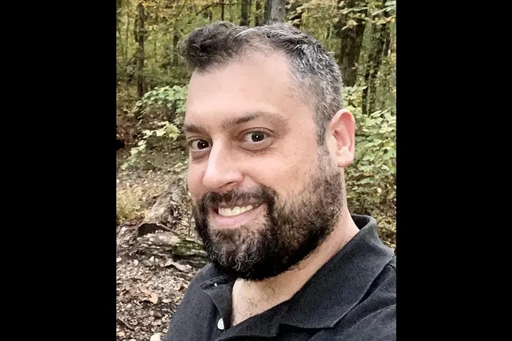 Poet Joshua St. Claire is standing in a forest. He is smiling with a brown beard.