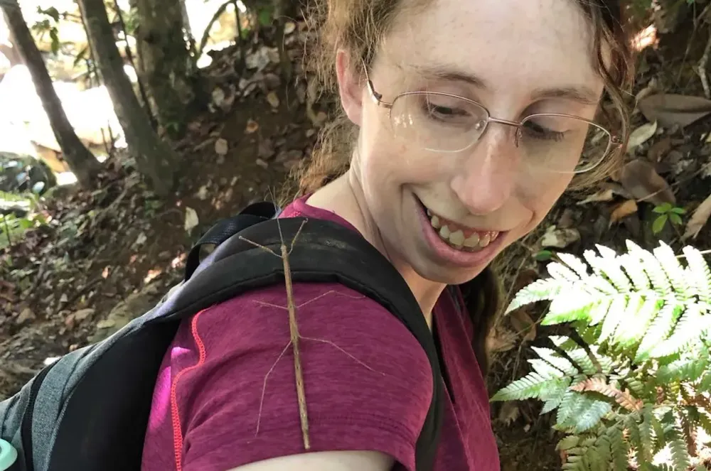 Pale freckled woman with wavy tied-back hair and a pinkish shirt and backpack on smiling at a walking stick that is crawling on her shoulder, a tropical forest is visible in the background.