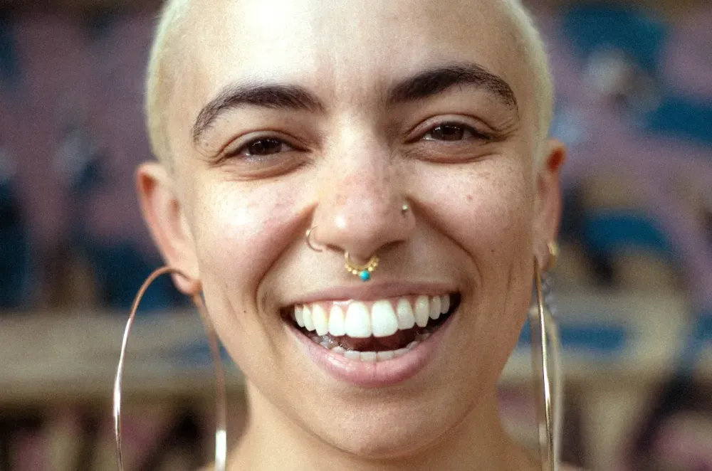 An androgynous person with bleached, shaved hair and dark eyebrows smiles widely. They are wearing large gold hoops and a necklace that reads Auslander.