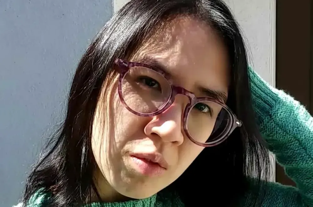 Susan is outdoors; the sun is shining on her face. She has shoulder-length black hair and wears glasses with purple tortoiseshell frames.