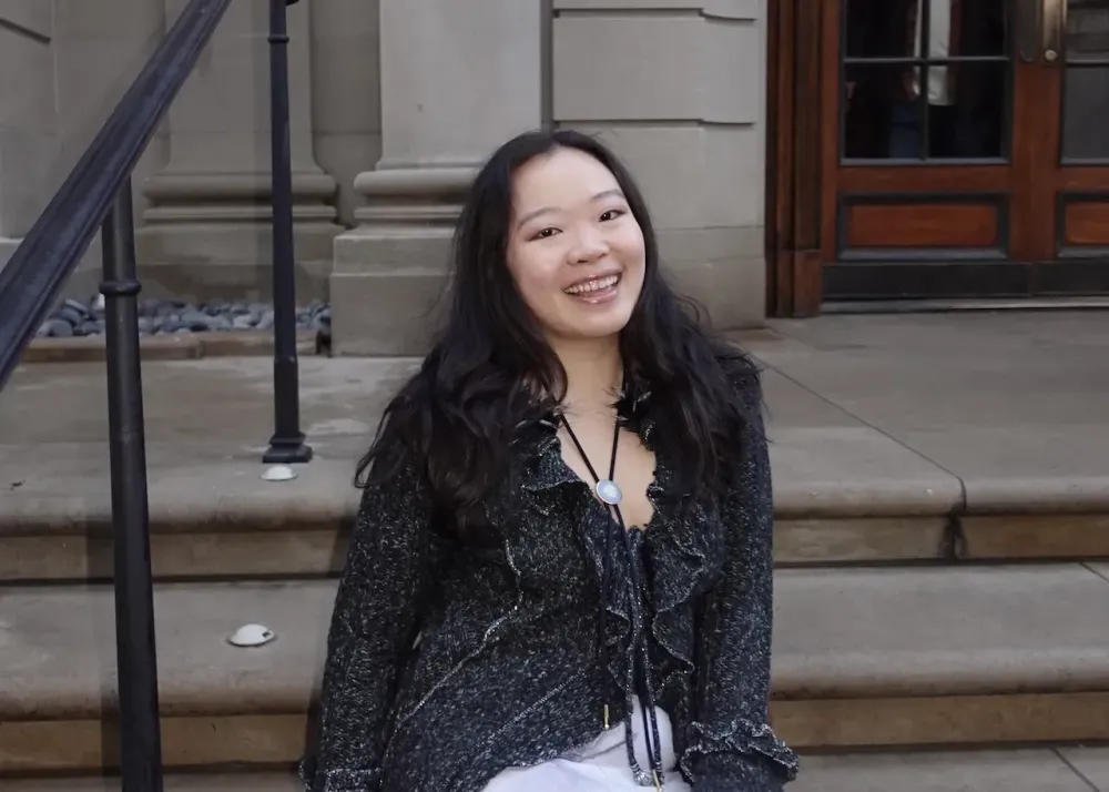 The author, Jessica Bao, Asian woman with black hair, sitting outside on the staircase at the entrance to the The Frick Collection in New York City.