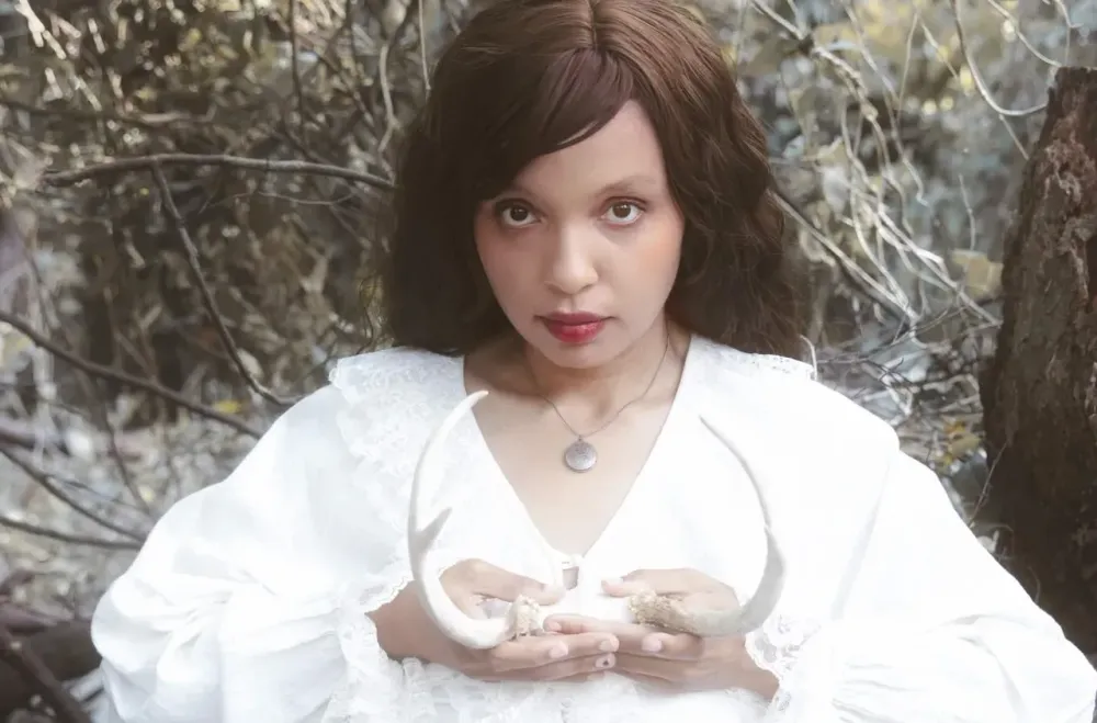 A person in the forest in a white dress holding antlers.