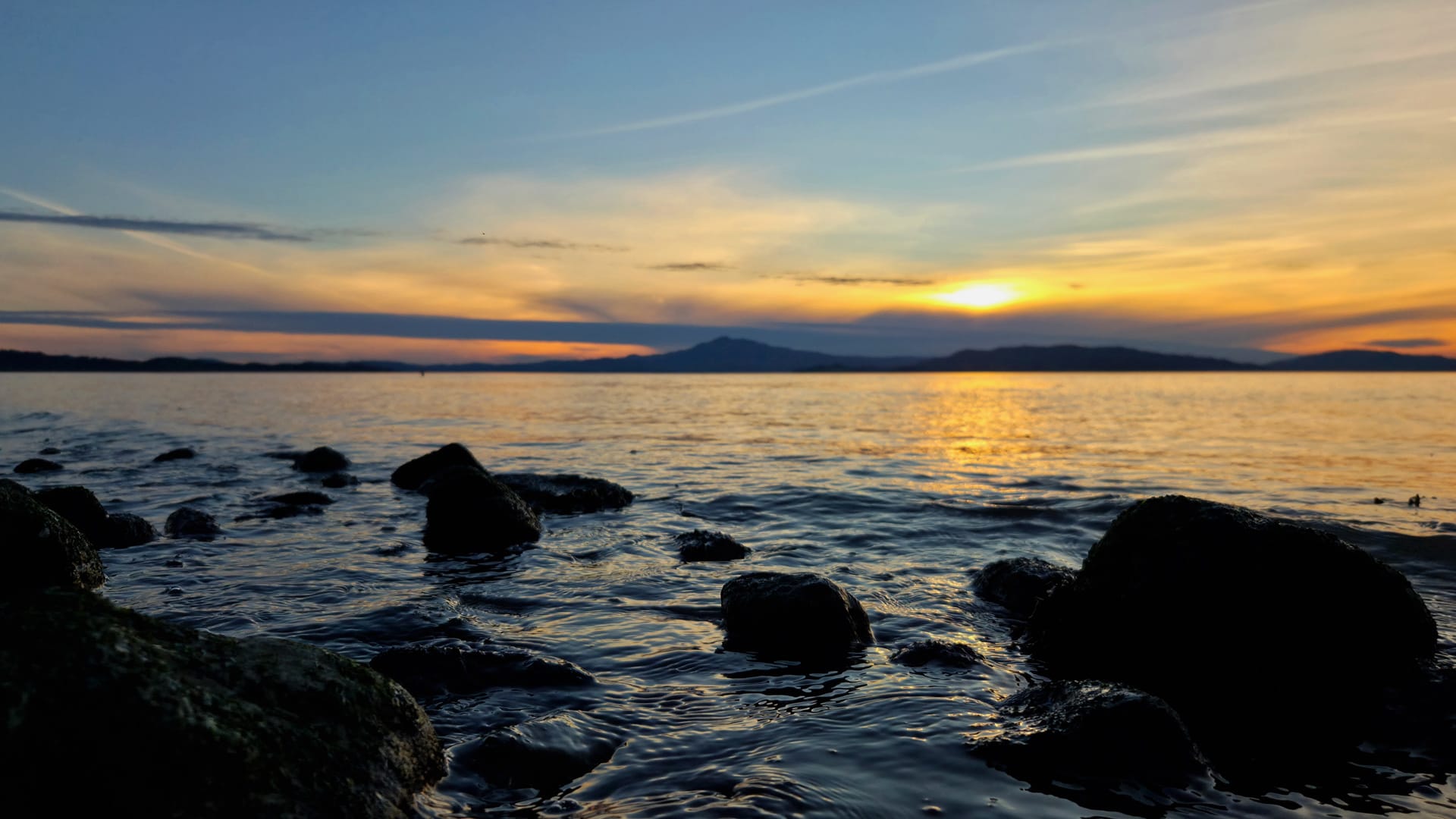 Rocky shoreline at blue hour, warm sunset light reflecting across the bay, Mt. Tamalpais silhouetted against a sky transitioning from deep orange at the horizon to blue overhead, moss-covered rocks in the foreground.
