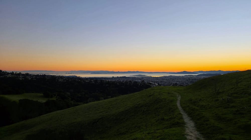 Sunset view from an East Bay trail looking across the bay toward the San Francisco skyline, warm light on the water, distant ridgeline fading into haze.