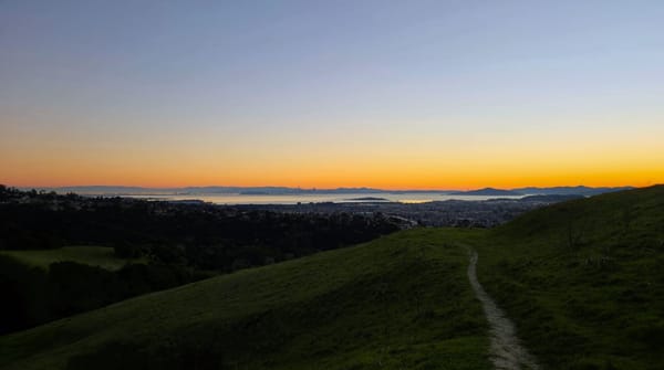 Sunset view from an East Bay trail looking across the bay toward the San Francisco skyline, warm light on the water, distant ridgeline fading into haze.
