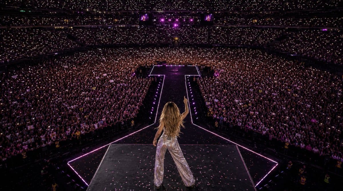 Female performer in sequined outfit on a dark stage in a packed stadium, viewed from behind with arms raised amid a massive crowd and purple lights.