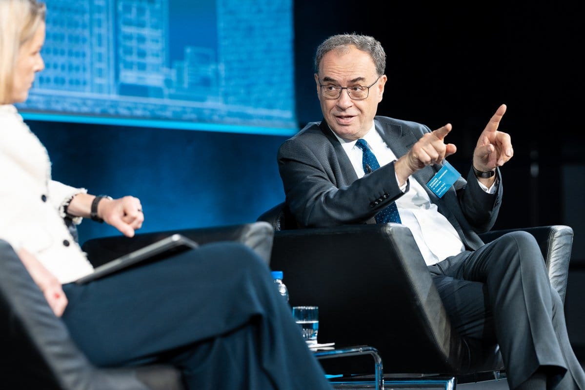 Andrew Bailey in a suit gesturing with both hands while seated at a conference next to a woman in a white jacket, with a blue city skyline screen behind them.