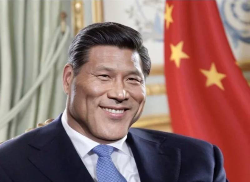 Close-up portrait of a smiling man in a dark suit and blue tie with the Chinese flag in the background.